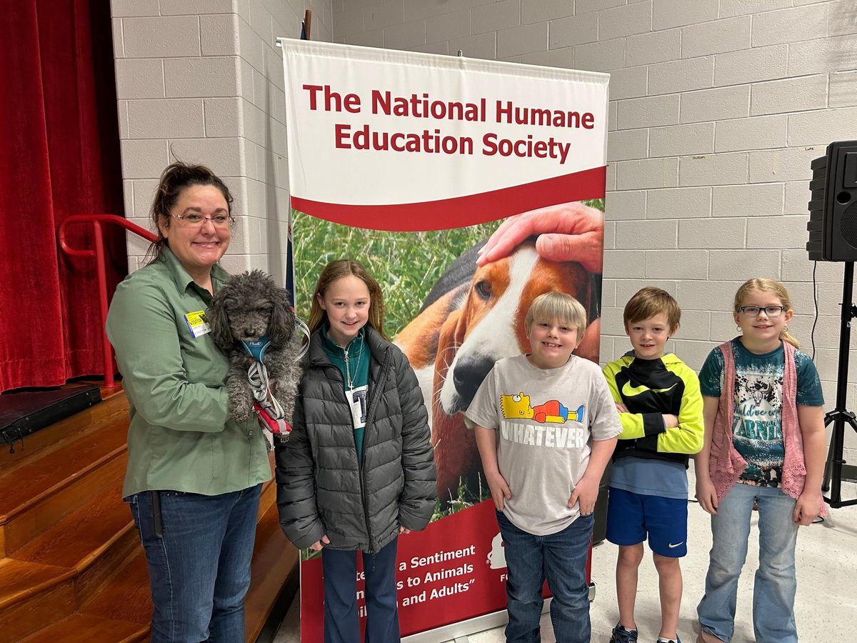 An NHES humane educator holding a small dog with four students in front of a National Humane Education Society banner